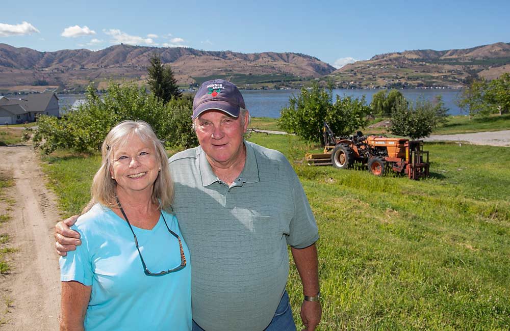 John and Sherry Kuntz pose in front of the site of their third-generation fruit orchard in August in Chelan, Washington. A year ago, the Kuntzes sold their share of the business to some nephews who may use it for wine grapes and vacation rentals and have been removing trees. The retired couple kept at least one tree of each variety for nostalgia’s sake. (Ross Courtney/Good Fruit Grower)