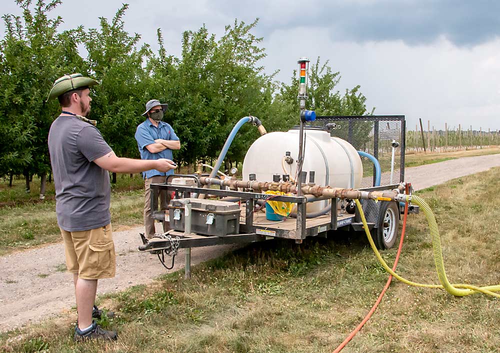 Project manager Keith Koonter, left, and Grieshop observe the linear version of SSCDS in action last July. Liquid is pumped from the spray tank to the orchard via the wider yellow pipe at right. The narrower yellow pipe is a return line. The orange line runs from a nearby air compressor. Gas powers the whole system. (Matt Milkovich/Good Fruit Grower)