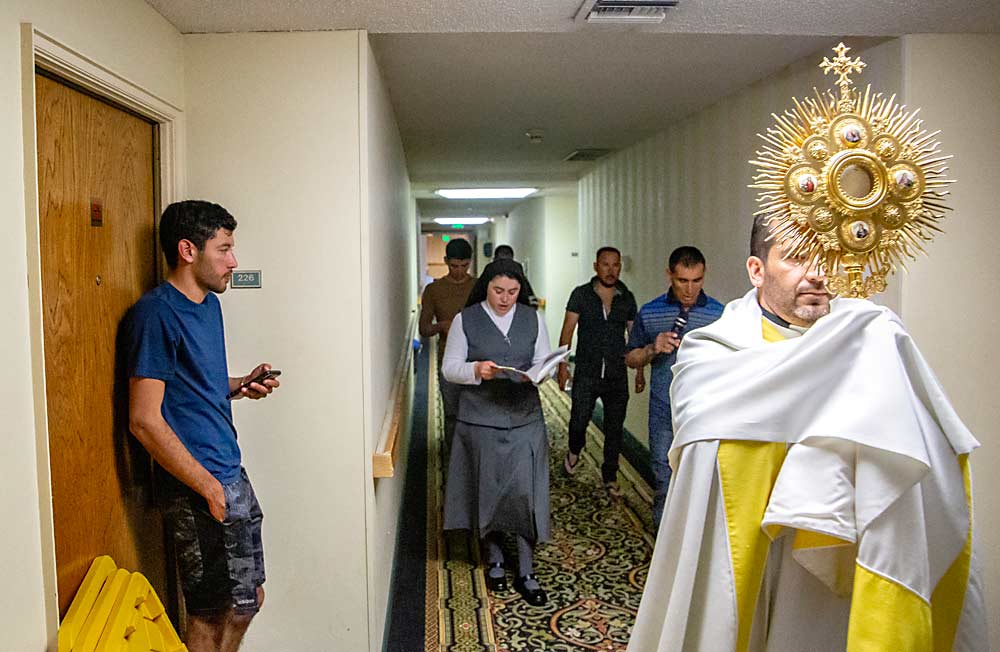 Father José De Jesús Mariscal Guzmán leads a worship processional in June through the hallways of a hotel used for H-2A housing in Yakima, Washington, while laborer Jesus Gutierrez Colima, left, checks on the commotion outside his room. (Ross Courtney/Good Fruit Grower)