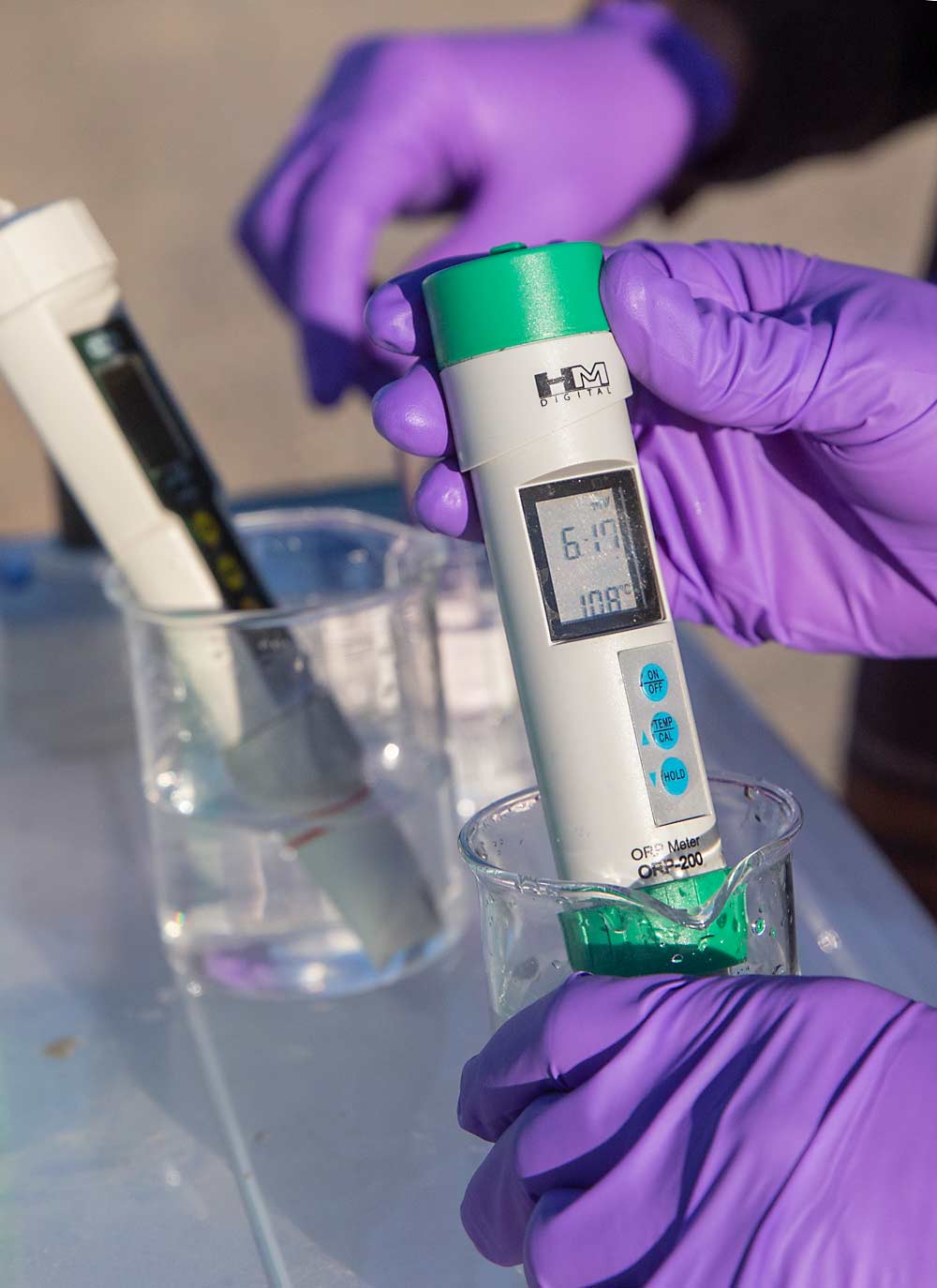 Anura Rathnayake-Mudiyanselage, a postdoctoral researcher, measures oxygen reduction potential and temperature of the ozonated water in the sprayer, while doctoral student Jake Schrader tests ozone concentration in the background. (Ross Courtney/Good Fruit Grower)