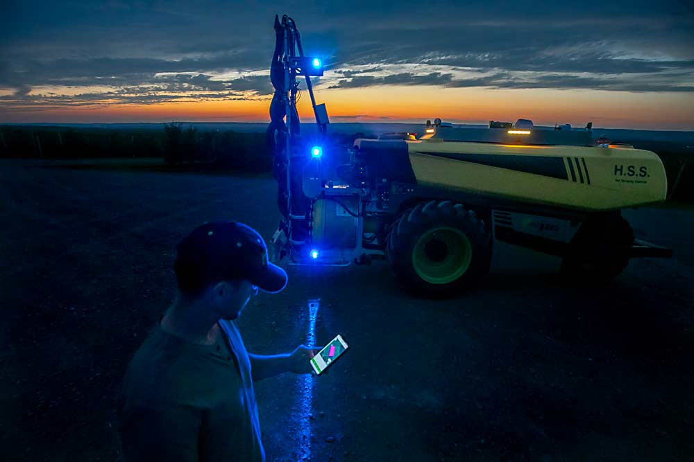 Chris Bartlett, from Provide Agro, operates the autonomous AgBot sprayer from his phone, sending it on a preplanned route at sunset through Crisp Growers’ high-density trellised orchards near Berwick, Nova Scotia, in July. (TJ Mullinax/Good Fruit Grower)